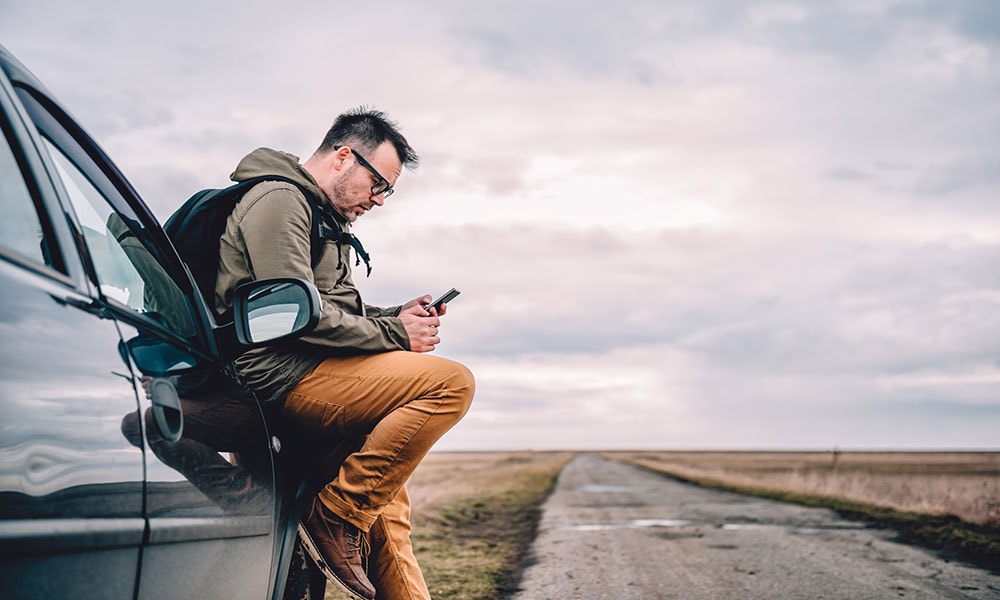 A man with a beard checking his phone while leaning against his car parked on the side of a country road in a remote location.