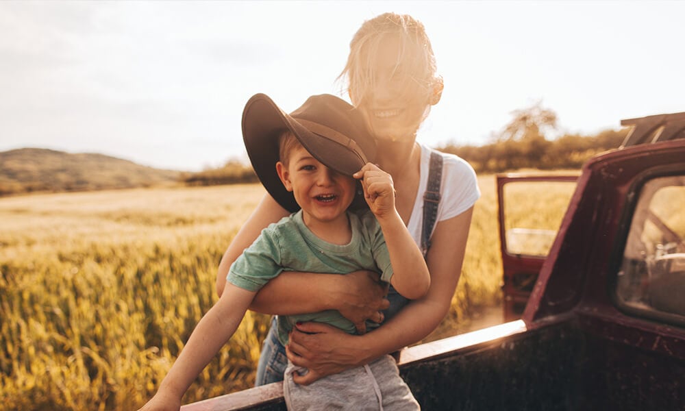 A young woman holds a young boy who is standing in the back of a utility vehicle in a rural location.
