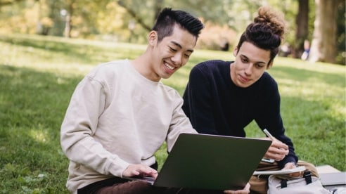 two male students sitting on grass and looking at a laptop